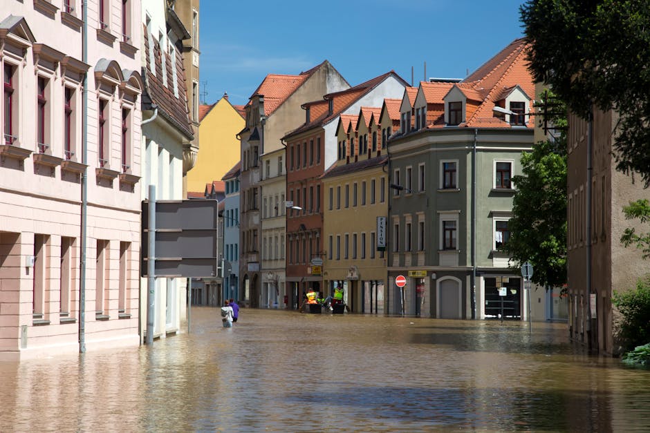 A street scene in Norwood Green showing several multi-story buildings with pastel-colored facades—pink, yellow, green, and beige—under a bright blue sky. Floodwaters have risen to cover the lower 