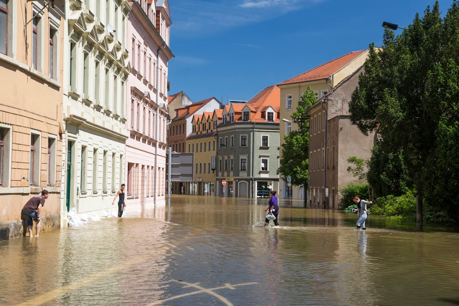 A street scene in Norwood Green showing several multi-story buildings with pastel-colored facades—pink, yellow, green, and beige—under a bright blue sky. Floodwaters have risen to cover the lower 