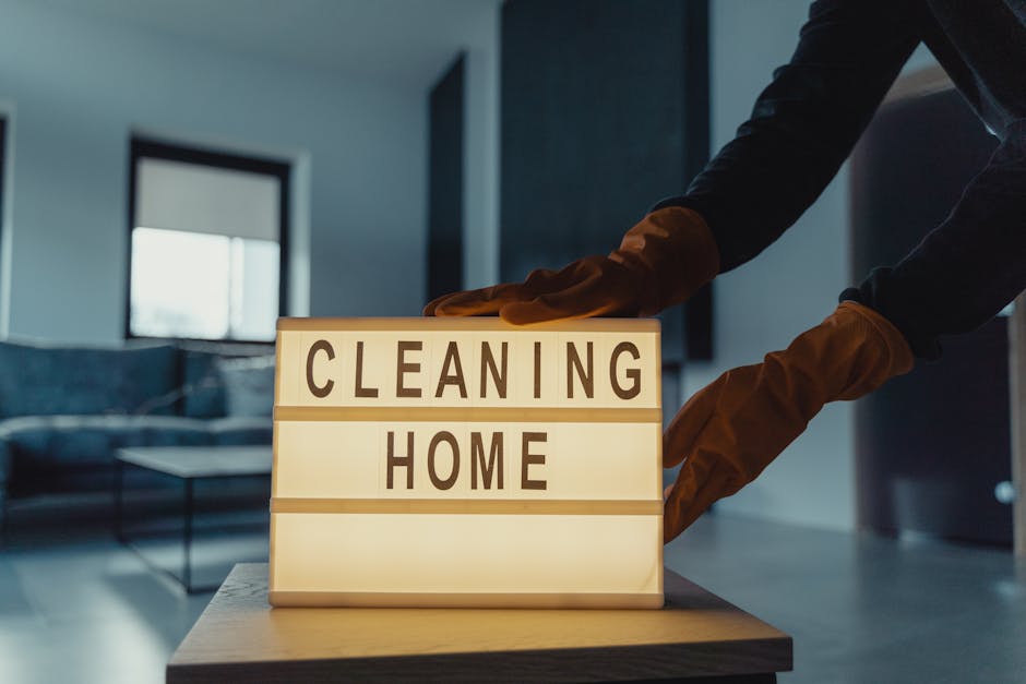 Close-up of a person wearing an orange glove, dusting or wiping a lightbox sign that reads 'CLEANING HOME' in bold black letters, placed on a wooden table. The background features a modern, well-lit r