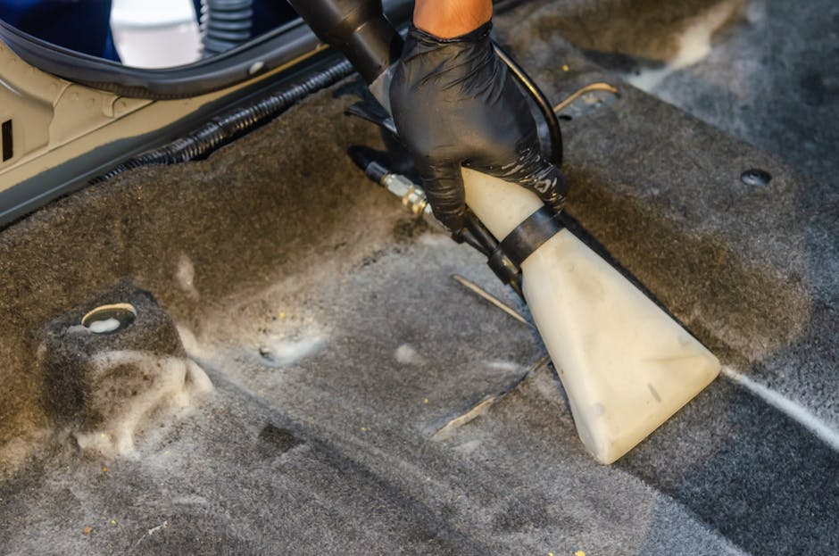 A person using a handheld vacuum cleaner to deep clean a yellow and white patterned cushion on a dark fabric sofa inside a domestic living room. The vacuum nozzle is pressed onto the cushion, which ap